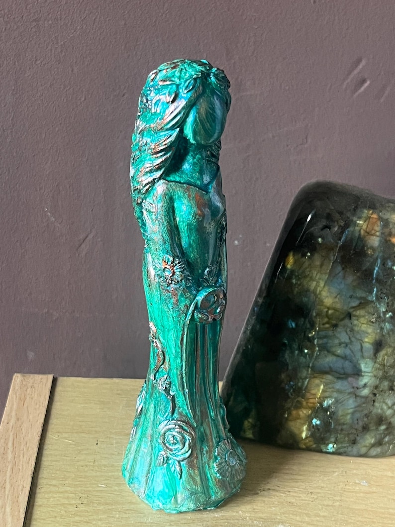 Puede incluir: Una estatua de color turquesa de una mujer con un vestido fluido, adornada con detalles florales. La estatua tiene una apariencia envejecida y antigua, con detalles en bronce. La figura est&aacute; junto a una gran piedra iridiscente.