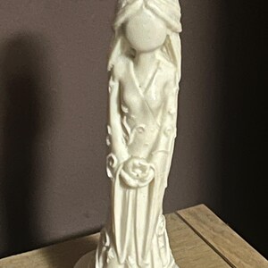 Puede incluir: Figurilla de color marfil de una mujer con un vestido largo, sosteniendo una rosa. La estatua tiene un acabado liso y detalles florales en el vestido y la base. La cara de la mujer es lisa, sin rasgos.