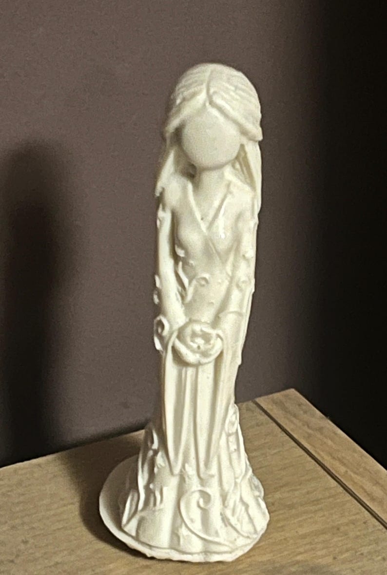 Puede incluir: Figurilla de color marfil de una mujer con un vestido largo, sosteniendo una rosa. La estatua tiene un acabado liso y detalles florales en el vestido y la base. La cara de la mujer es lisa, sin rasgos.