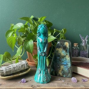 Puede incluir: Una estatua turquesa de una mujer sosteniendo una luna creciente, con detalles florales. Una piedra grande e iridiscente y un manojo de salvia descansan sobre una superficie de madera, con una planta verde en el fondo.