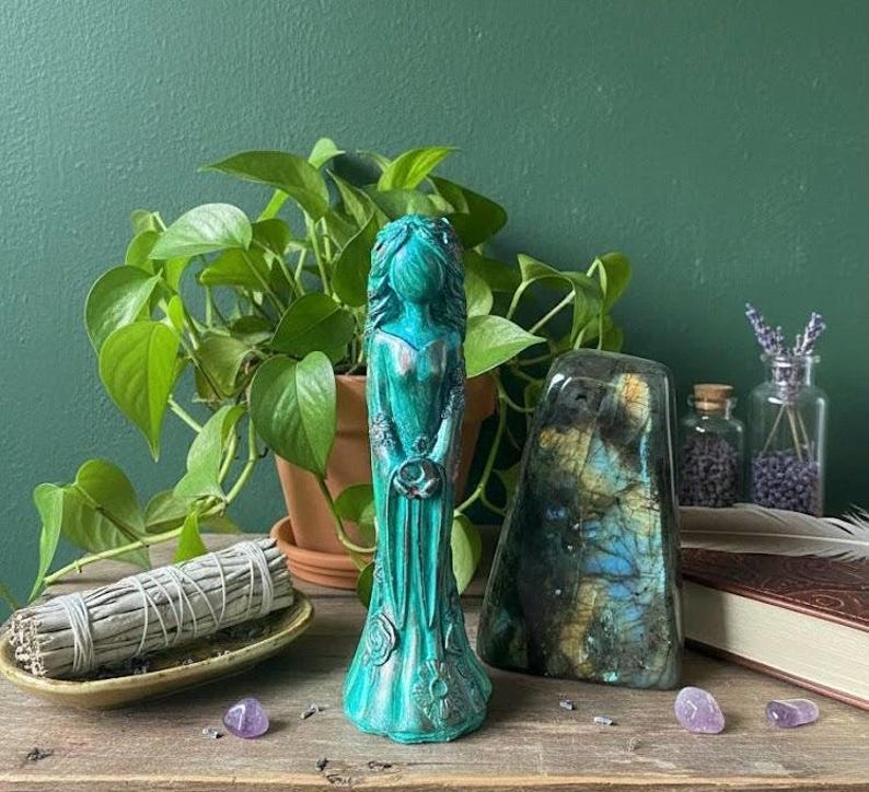 Puede incluir: Una estatua turquesa de una mujer sosteniendo una luna creciente, con detalles florales. Una piedra grande e iridiscente y un manojo de salvia descansan sobre una superficie de madera, con una planta verde en el fondo.