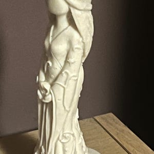 Puede incluir: Figura de color crema de una mujer con un vestido fluido. La estatua tiene pelo largo y sostiene un objeto peque&ntilde;o. El vestido presenta detalles florales y de vid. La figura est&aacute; sobre una superficie de madera.