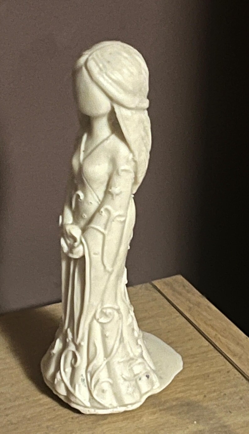 Puede incluir: Figura de color crema de una mujer con un vestido fluido. La estatua tiene pelo largo y sostiene un objeto peque&ntilde;o. El vestido presenta detalles florales y de vid. La figura est&aacute; sobre una superficie de madera.