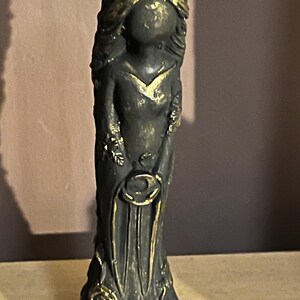 Puede incluir: Una estatua gris oscuro de una mujer con un vestido largo, adornada con detalles dorados. La estatua presenta una corona floral, un escote en V y dise&ntilde;os florales y circulares. La est&eacute;tica general recuerda al arte antiguo.