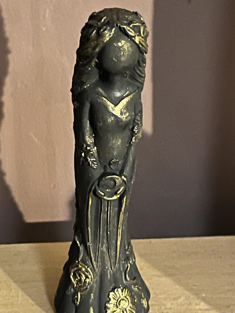 Puede incluir: Una estatua gris oscuro de una mujer con un vestido largo, adornada con detalles dorados. La estatua presenta una corona floral, un escote en V y dise&ntilde;os florales y circulares. La est&eacute;tica general recuerda al arte antiguo.