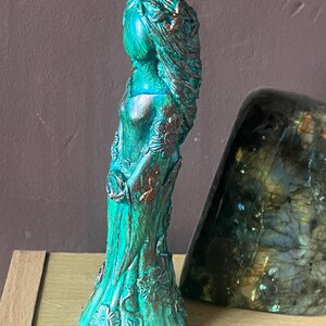 Puede incluir: Figura turquesa de una mujer con pelo largo y vestido fluido, adornada con detalles florales y acentos de bronce. La estatua est&aacute; sobre una superficie de madera.