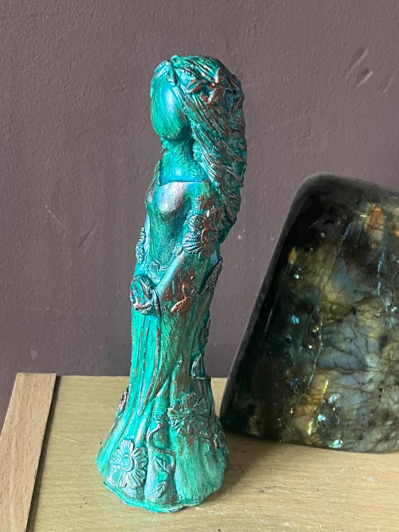 Puede incluir: Figura turquesa de una mujer con pelo largo y vestido fluido, adornada con detalles florales y acentos de bronce. La estatua est&aacute; sobre una superficie de madera.