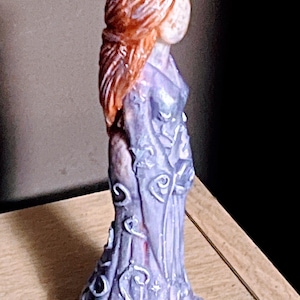Puede incluir: Peque&ntilde;a figura decorativa de una mujer con un vestido largo y fluido. La figura tiene cabello rojo-anaranjado y una cara p&aacute;lida. El vestido es de color lila claro con detalles florales blancos. La base es blanca.