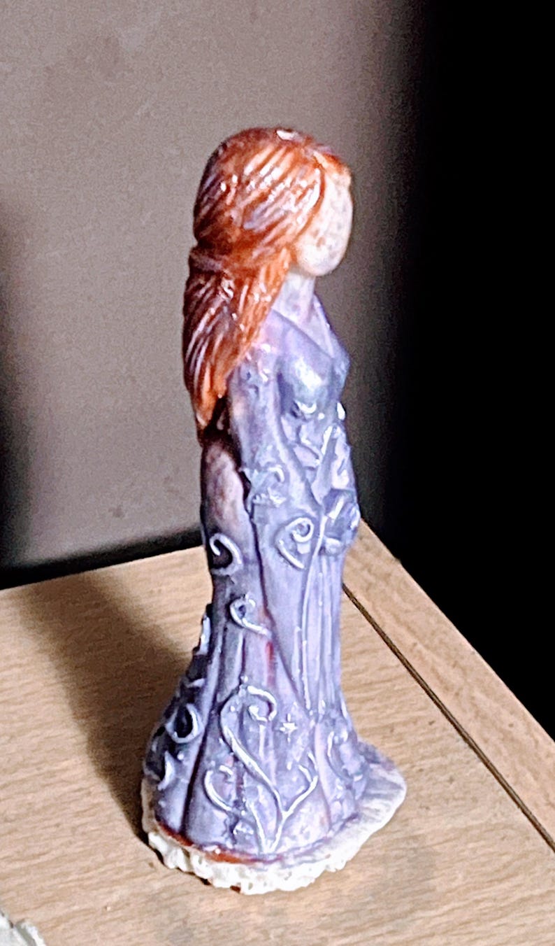 Puede incluir: Peque&ntilde;a figura decorativa de una mujer con un vestido largo y fluido. La figura tiene cabello rojo-anaranjado y una cara p&aacute;lida. El vestido es de color lila claro con detalles florales blancos. La base es blanca.