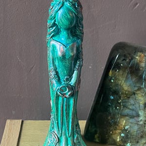 Puede incluir: Una estatua de color turquesa de una mujer con cabello largo y suelto y un vestido largo. La estatua tiene detalles en color bronce y detalles florales. La estatua est&aacute; sobre una superficie de color claro.