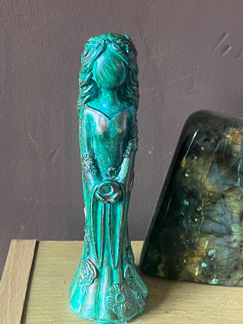 Puede incluir: Una estatua de color turquesa de una mujer con cabello largo y suelto y un vestido largo. La estatua tiene detalles en color bronce y detalles florales. La estatua est&aacute; sobre una superficie de color claro.