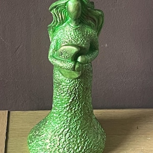 Pode incluir: Estátua verde de uma figura com cabelo esvoaçante, segurando uma forma de lua crescente com uma estrela. A estátua tem uma superfície texturizada e é pintada em verde metálico. A figura está vestindo um vestido longo.