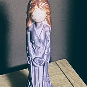 Puede incluir: Una figura de color lavanda de una mujer con un vestido fluido. La estatua tiene cabello casta&ntilde;o rojizo y sostiene un objeto peque&ntilde;o. El vestido presenta intrincados patrones florales y un dobladillo decorativo. La figura est&aacute; sobre una superficie de madera.