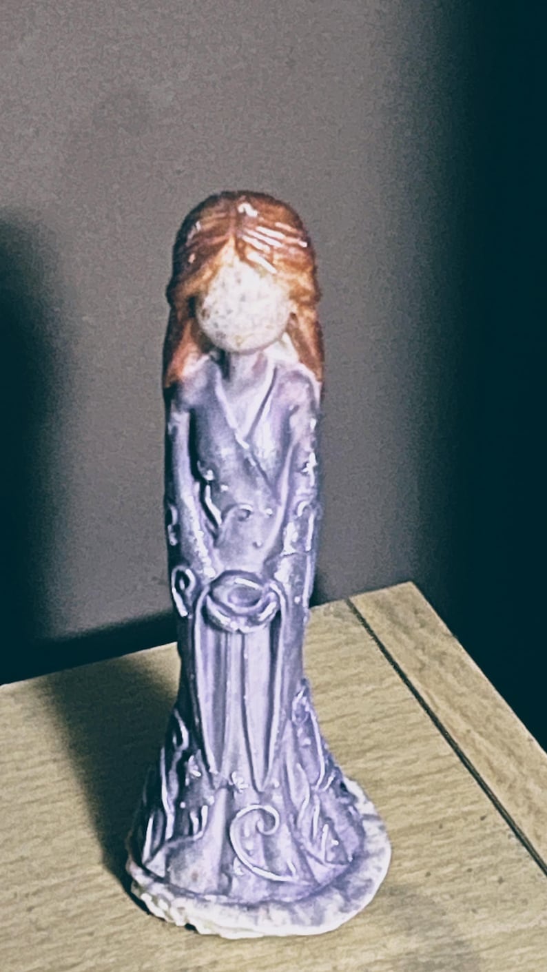 Puede incluir: Una figura de color lavanda de una mujer con un vestido fluido. La estatua tiene cabello casta&ntilde;o rojizo y sostiene un objeto peque&ntilde;o. El vestido presenta intrincados patrones florales y un dobladillo decorativo. La figura est&aacute; sobre una superficie de madera.