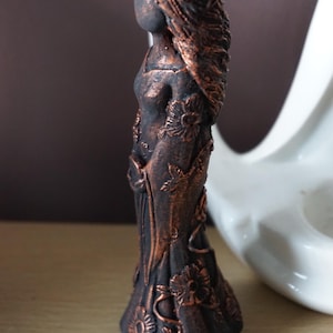 Puede incluir: Estatua de bronce de una mujer con pelo largo y una corona floral. La figura est&aacute; adornada con motivos florales en su vestido. La estatua mide aproximadamente 15 cm de altura y se encuentra sobre una superficie de madera.