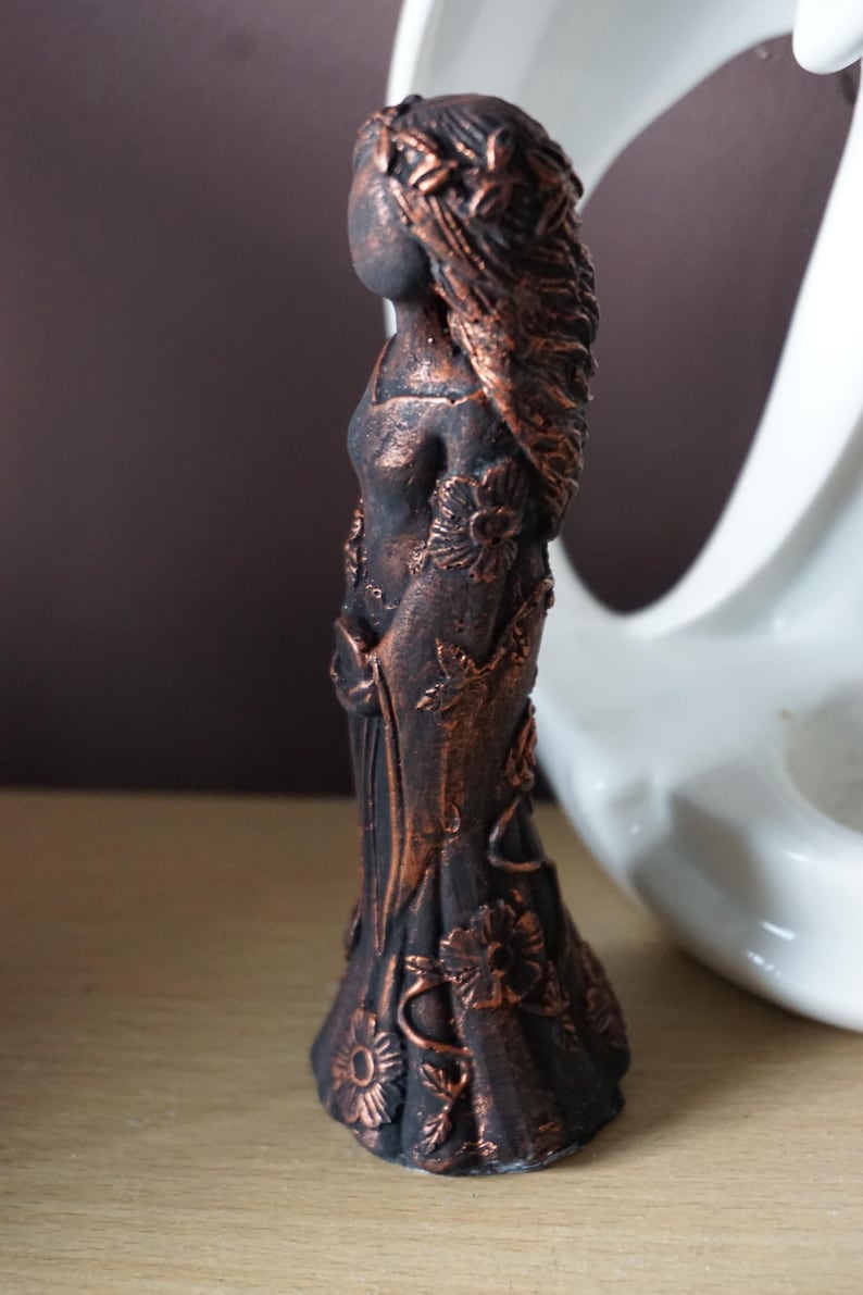 Puede incluir: Estatua de bronce de una mujer con pelo largo y una corona floral. La figura est&aacute; adornada con motivos florales en su vestido. La estatua mide aproximadamente 15 cm de altura y se encuentra sobre una superficie de madera.