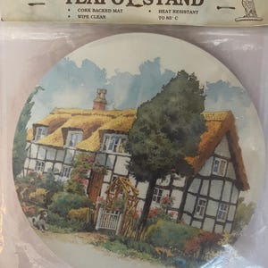 Könnte beinhalten: Ein runder Teekannenuntersetzer in Kunststoffverpackung mit einer Aquarellillustration eines strohgedeckten Hauses mit Fachwerk, einem Garten und einem kleinen Hund. Der Text auf der Verpackung lautet SE Giftware Manufacturers, TEAPOT STAND, CORK BACKED MAT, WIPE CLEAN und HEAT RESISTANT TO 85 C.