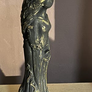 Puede incluir: Una estatua gris oscuro de una mujer con un vestido fluido, adornada con detalles florales dorados. La estatua presenta detalles intrincados, incluyendo una corona floral y delicados adornos en el vestido. La figura est&aacute; de perfil, mostrando el elegante dise&ntilde;o.
