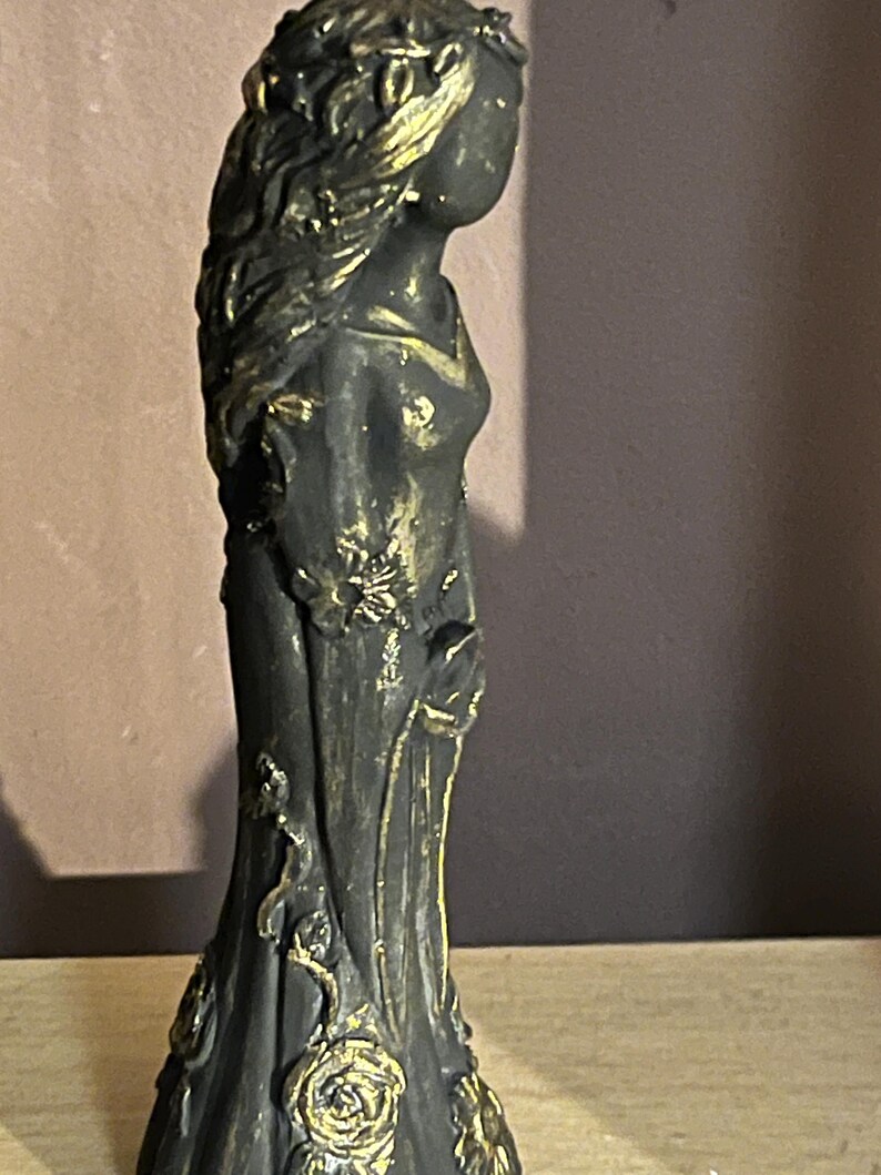 Puede incluir: Una estatua gris oscuro de una mujer con un vestido fluido, adornada con detalles florales dorados. La estatua presenta detalles intrincados, incluyendo una corona floral y delicados adornos en el vestido. La figura est&aacute; de perfil, mostrando el elegante dise&ntilde;o.