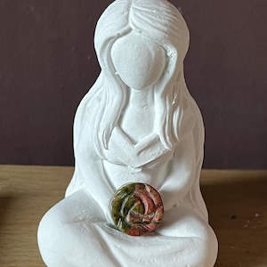 Peut inclure: Figurines en céramique blanche d'une femme assise les jambes croisées, tenant une pierre ronde verte, rose et brune sur ses genoux.