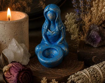 Puede incluir: Una figura azul de una silueta sentada con cabello largo, que presenta una hendidura cóncava en forma de cuenco en el regazo. La escultura tiene un acabado texturizado con sutiles reflejos dorados en los bordes y contornos.