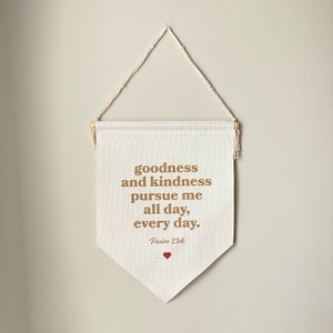 May include: A cream-colored banner with the text "goodness and kindness pursue me all day, every day." in a brown font. The banner also includes the text "Psalm 23:6" and a small red heart. The banner is suspended by a wooden rod and a cream-colored string.