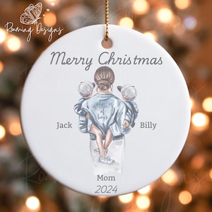 Könnte beinhalten: Weißer Keramik-Weihnachtsbaumschmuck mit einer Aquarell-Illustration einer Frau mit zwei Jungen. Die Frau trägt eine blaue Jeansjacke mit dem Schriftzug "Mom of boys" auf dem Rücken. Die Jungen tragen blaue Baseballkappen. Der Text "Merry Christmas" befindet sich oben auf dem Ornament. Der Text "Jack" und "Billy" befinden sich auf beiden Seiten der Frau. Der Text "Mom 2024" befindet sich unten auf dem Ornament.