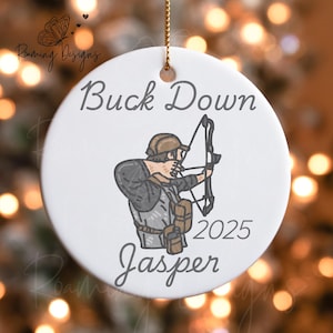 Peut inclure: Ornement de Noël rond en céramique blanche avec un dessin d'archer tirant à l'arc. Le texte "Buck Down 2025 Jasper" est écrit sur l'ornement. Une ficelle dorée est attachée pour l'accrocher.