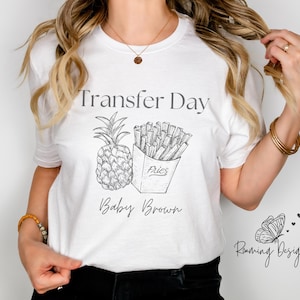 Puede incluir: Camiseta blanca con el texto "Transfer Day" y un dibujo de una piña y patatas fritas en una caja que dice "Fries". El texto "Baby Brown" está debajo del dibujo. La esquina inferior derecha de la camiseta tiene el texto "Running Designs" y una mariposa.