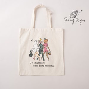 Puede incluir: Una bolsa de tela blanca con un diseño de temática de Halloween que presenta a tres mujeres vestidas como un fantasma, un esqueleto y una calabaza. El texto en la bolsa dice "Get in ghoulies, We're going haunting."