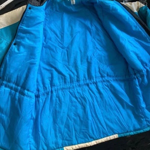 May include: A blue, white, and black puffer jacket with a zipper closure and a drawstring waist. The jacket is unzipped and the lining is visible.