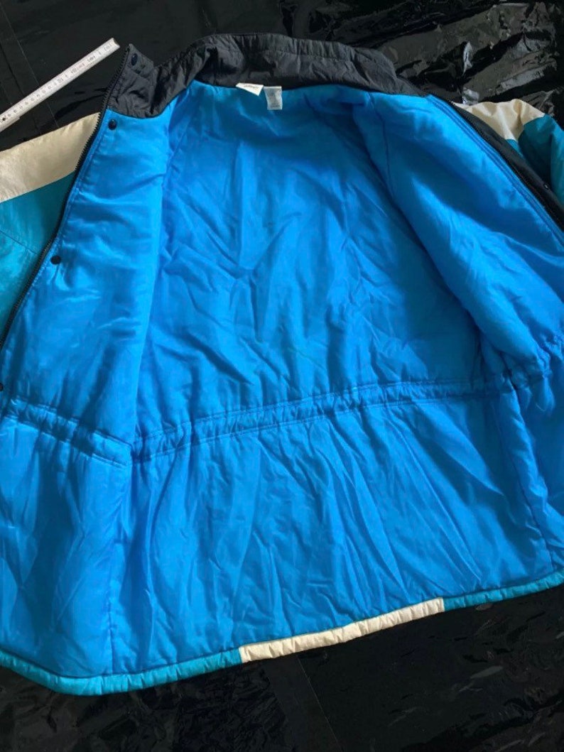 May include: A blue, white, and black puffer jacket with a zipper closure and a drawstring waist. The jacket is unzipped and the lining is visible.