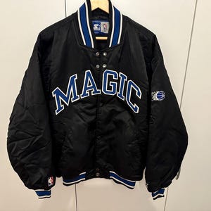 Könnte beinhalten: Schwarze Satinjacke mit dem Wort "MAGIC" in Weiß und Blau auf der Brust. Die Jacke hat einen blau-weiß gestreiften Kragen, Bündchen und Saum. Ein Basketball-Logo befindet sich auf dem Ärmel.