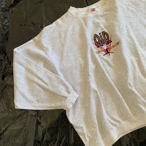 K&ouml;nnte beinhalten: Grauer Sweatshirt mit einem gro&szlig;en gestickten Motiv eines Basketballspielers mit dem Wort "AIR" in orangefarbenen und violetten Buchstaben.