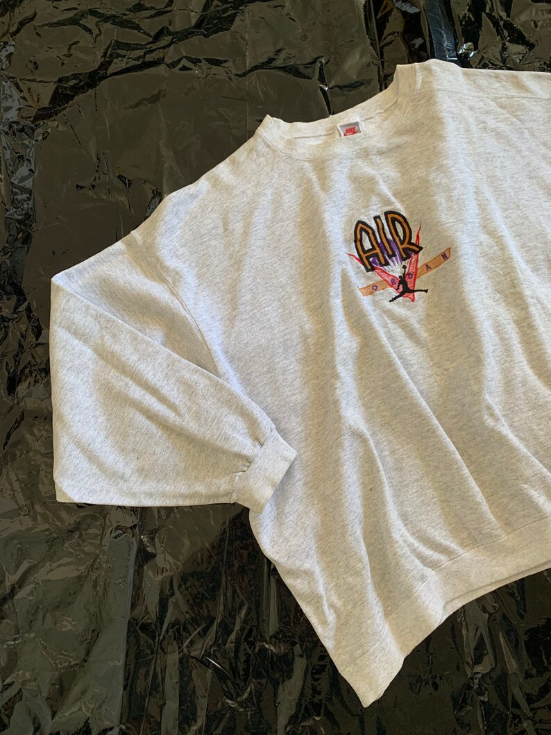 K&ouml;nnte beinhalten: Grauer Sweatshirt mit einem gro&szlig;en gestickten Motiv eines Basketballspielers mit dem Wort "AIR" in orangefarbenen und violetten Buchstaben.