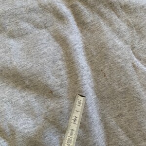 K&ouml;nnte beinhalten: Nahaufnahme einer grauen Stoffoberfl&auml;che mit zwei kleinen braunen Flecken. Ein Lineal ist teilweise in der rechten unteren Ecke des Bildes sichtbar.