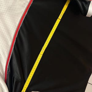 Peut inclure: Un maillot de sport noir et blanc avec une bordure rouge et grise. Un m&egrave;tre ruban jaune est pos&eacute; sur le maillot, indiquant une mesure d'environ 150 cm.