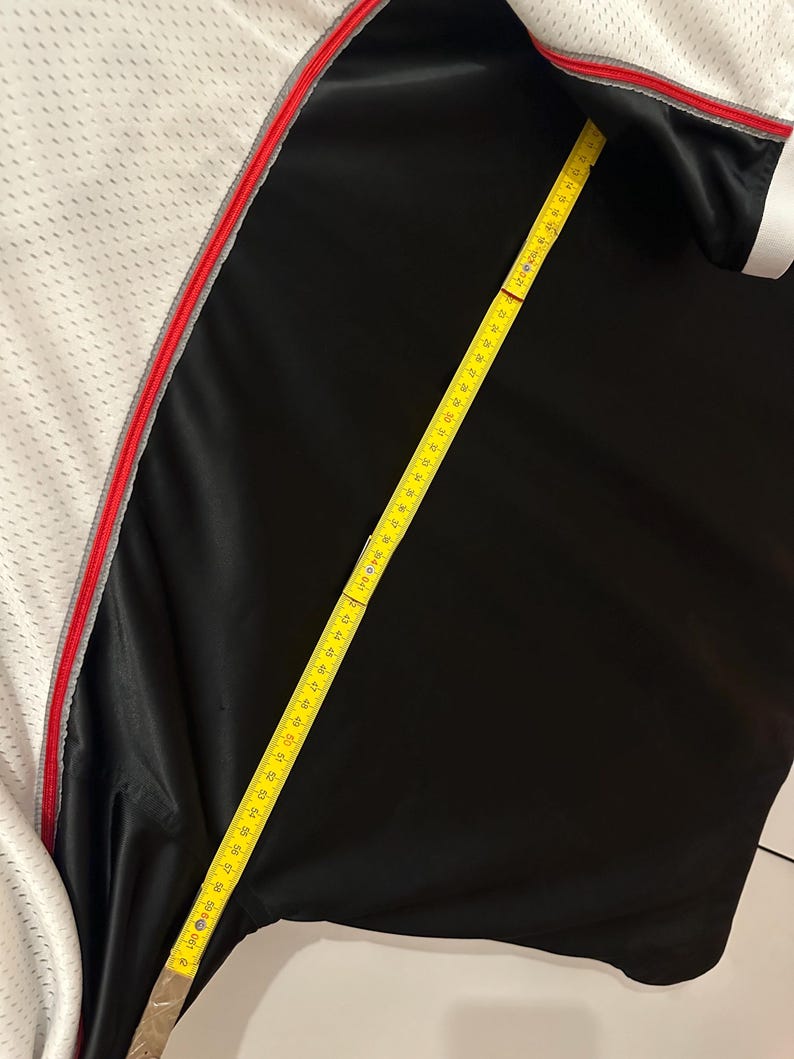 Peut inclure: Un maillot de sport noir et blanc avec une bordure rouge et grise. Un m&egrave;tre ruban jaune est pos&eacute; sur le maillot, indiquant une mesure d'environ 150 cm.