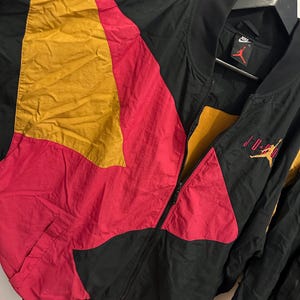 K&ouml;nnte beinhalten: Schwarze, rote und gelbe Jordan Bomberjacke mit durchgehendem Rei&szlig;verschluss. Die Jacke hat ein Colorblock-Design mit dem Jordan-Logo auf Brust und &Auml;rmel. Die Jacke h&auml;ngt an einem schwarzen Kleiderb&uuml;gel.