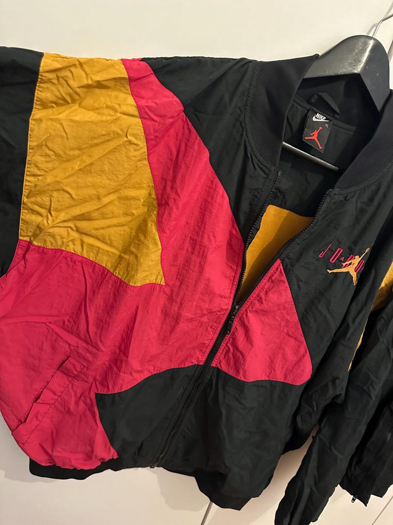 K&ouml;nnte beinhalten: Schwarze, rote und gelbe Jordan Bomberjacke mit durchgehendem Rei&szlig;verschluss. Die Jacke hat ein Colorblock-Design mit dem Jordan-Logo auf Brust und &Auml;rmel. Die Jacke h&auml;ngt an einem schwarzen Kleiderb&uuml;gel.