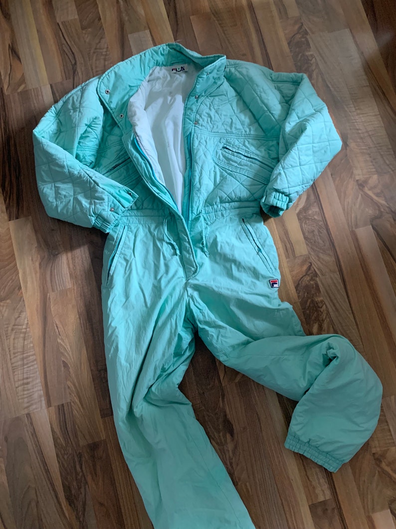 May include: A light blue ski suit with a quilted design and the word "FILA" on the chest.