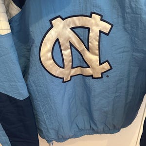 May include: Light blue and navy blue nylon jacket with a large, silver and black logo on the back. The jacket has a Starter logo on the lower left side. The jacket is a vintage style.