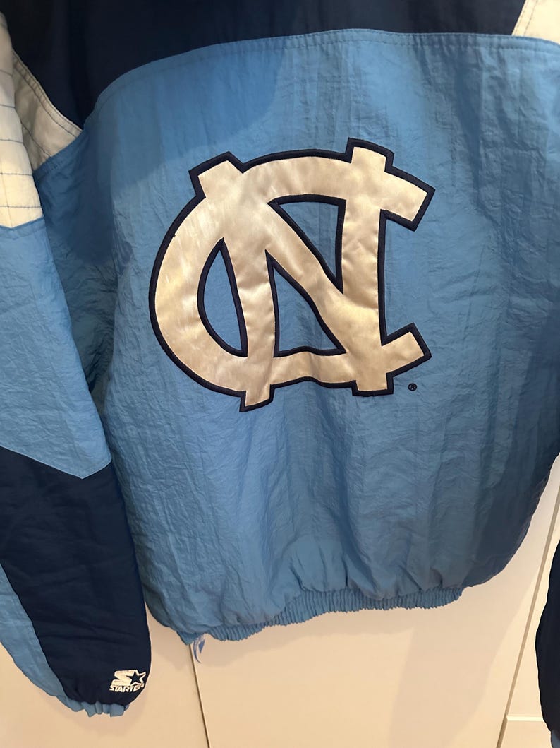 May include: Light blue and navy blue nylon jacket with a large, silver and black logo on the back. The jacket has a Starter logo on the lower left side. The jacket is a vintage style.