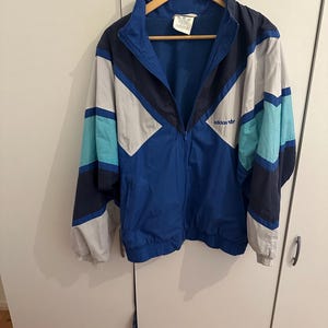 May include: A blue vintage Adidas windbreaker jacket with white, blue, and black color block design. The jacket has a zipper closure and a logo on the front that says "adidas".