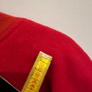 May include: A close-up of a red garment with a ribbed orange collar. A yellow ruler is placed diagonally across the garment, showing measurements in centimeters. The garment's texture appears soft and plush.