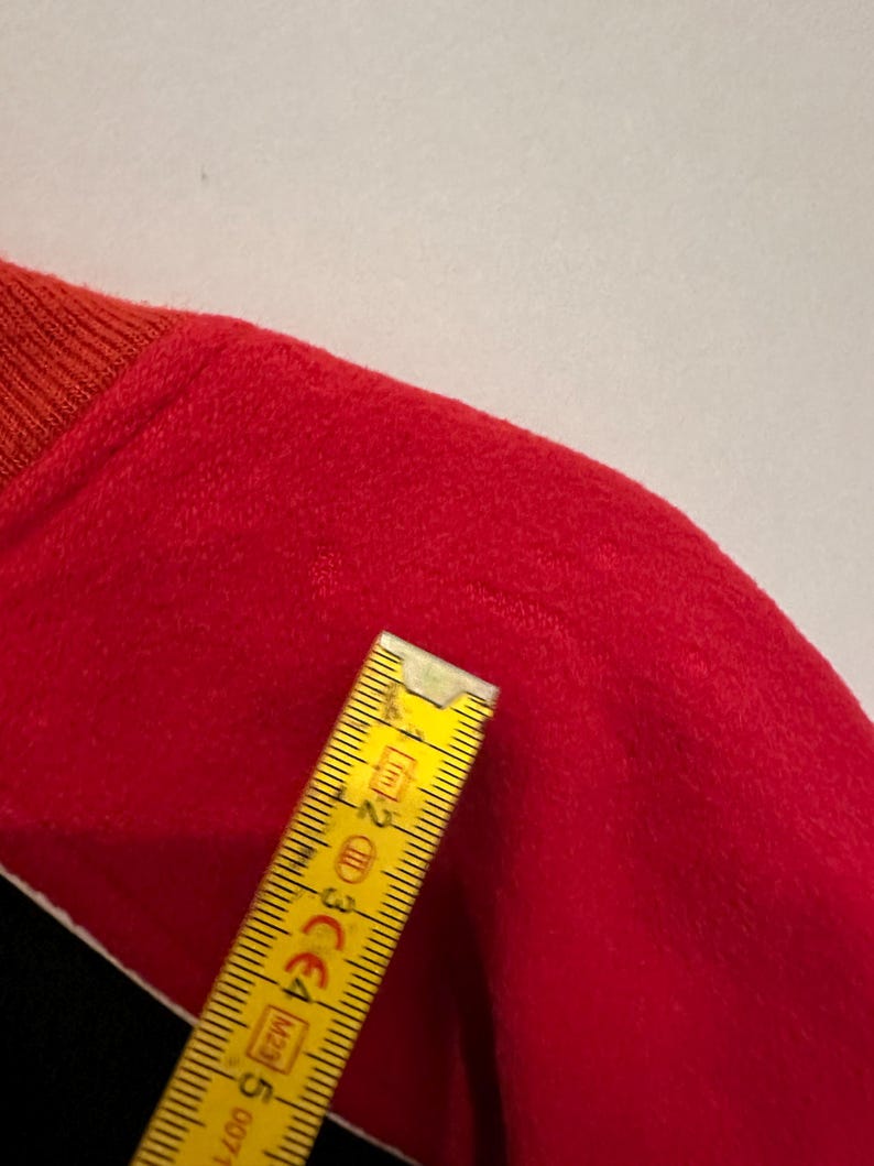 May include: A close-up of a red garment with a ribbed orange collar. A yellow ruler is placed diagonally across the garment, showing measurements in centimeters. The garment's texture appears soft and plush.
