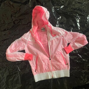 Könnte beinhalten: Eine rosafarbene Kapuzenjacke mit einem weißen Reißverschluss und weißen Besätzen. Die Jacke hat ein Nike-Logo auf der linken Brust.