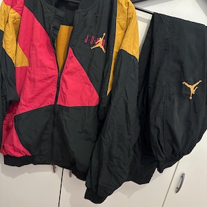 K&ouml;nnte beinhalten: Ein schwarzer, goldener und roter Jordan-Trainingsanzug. Die Jacke hat einen durchgehenden Rei&szlig;verschluss und das Jordan-Logo in Rot. Die Hose ist schwarz mit dem Jordan-Logo in Gold. Jacke und Hose h&auml;ngen an einem Kleiderb&uuml;gel.