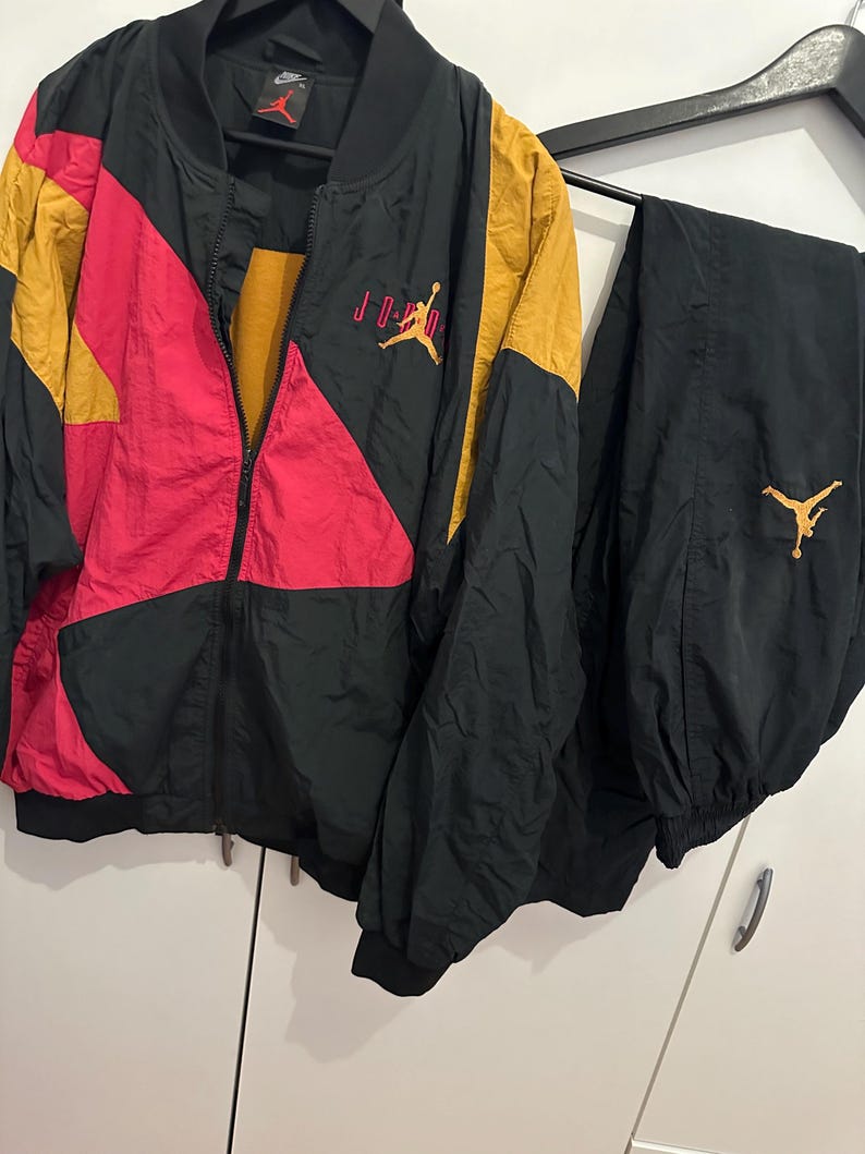K&ouml;nnte beinhalten: Ein schwarzer, goldener und roter Jordan-Trainingsanzug. Die Jacke hat einen durchgehenden Rei&szlig;verschluss und das Jordan-Logo in Rot. Die Hose ist schwarz mit dem Jordan-Logo in Gold. Jacke und Hose h&auml;ngen an einem Kleiderb&uuml;gel.
