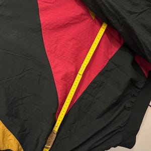 K&ouml;nnte beinhalten: Eine Vintage-Windjacke in Schwarz, Rot und Gold. Die Jacke wird mit einem gelben Ma&szlig;band gemessen. Die Windjacke hat ein Farbblock-Design mit einem geometrischen Muster. Die Jacke ist aus leichtem Material gefertigt.